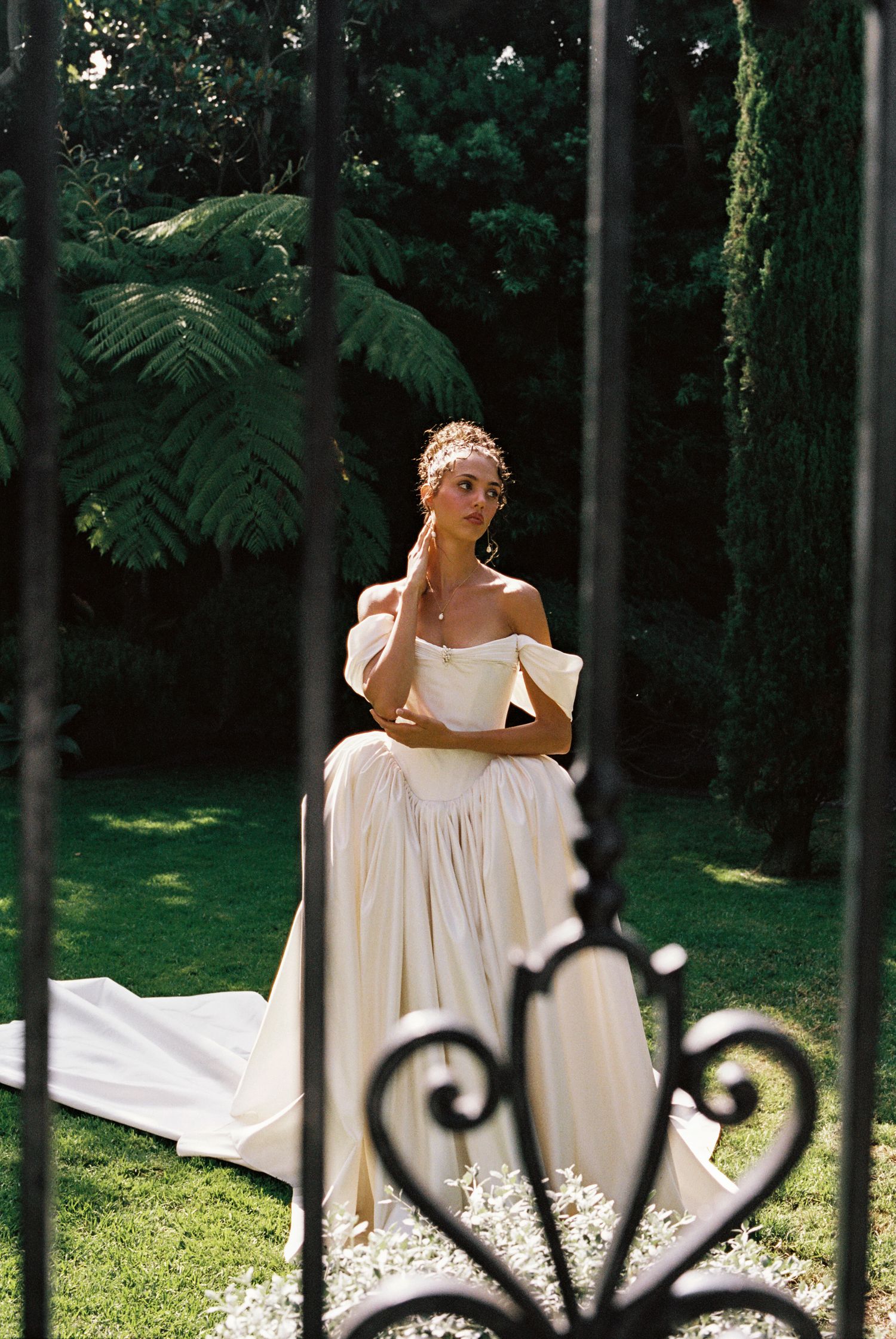 Bride in off-shoulder ivory gown with floral crown poses gracefully in lush garden, viewed through ornate iron gates.