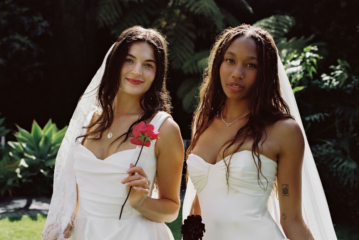 Two brides in white strapless gowns and veils pose outdoors, one holding a red rose, surrounded by lush greenery.