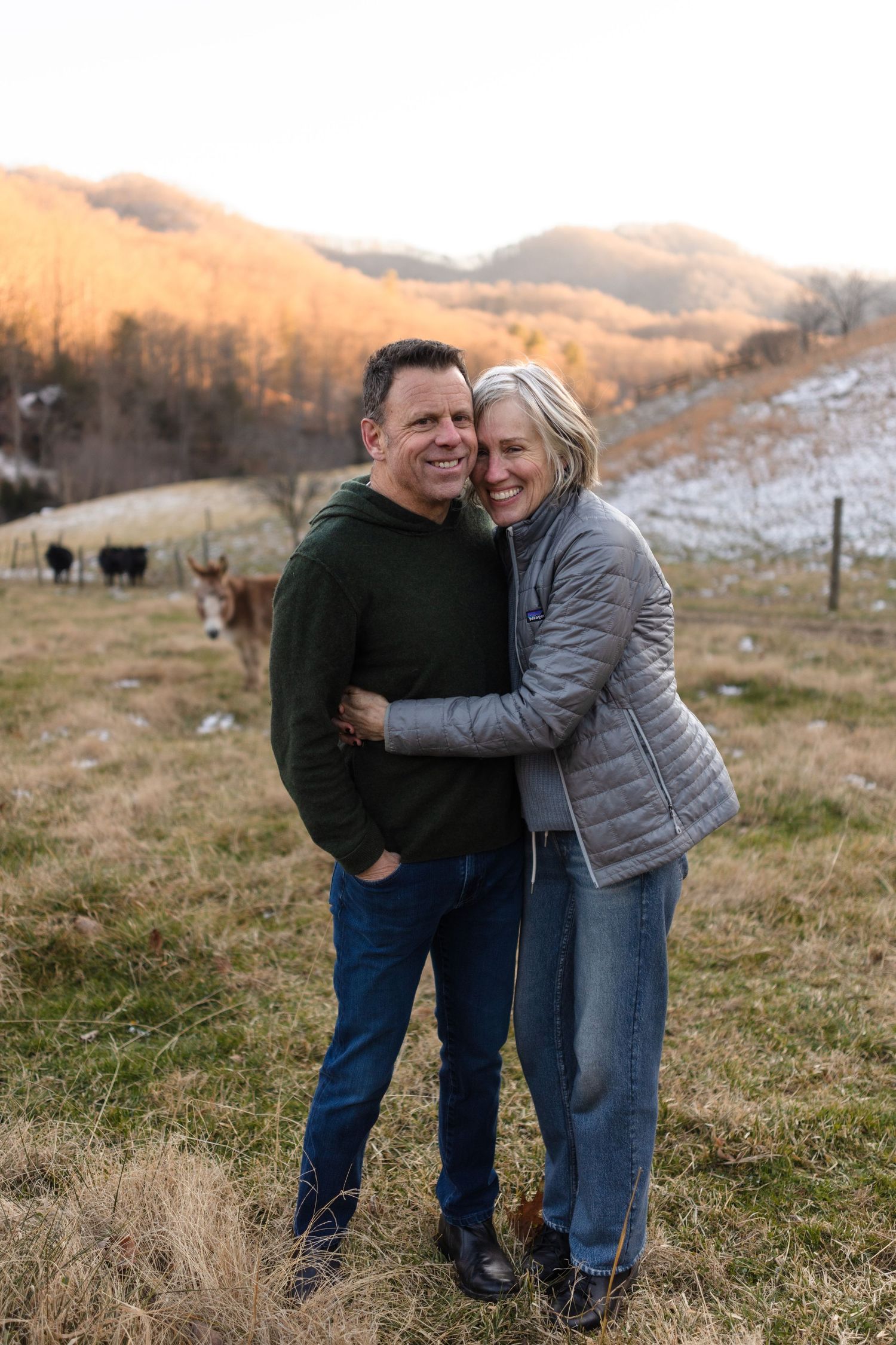 A smiling couple embraces in a rural mountain field at sunset, with cattle and snow-dusted hills in the background.