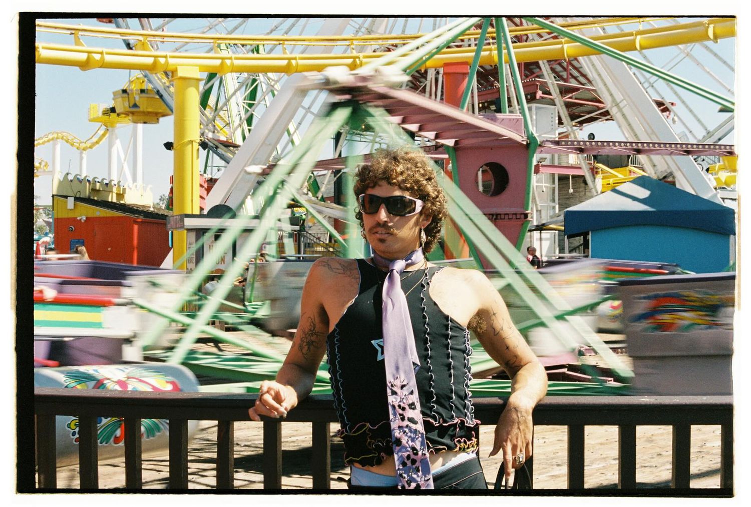 Person in black vest and tie standing before colorful amusement park rides on sunny day.