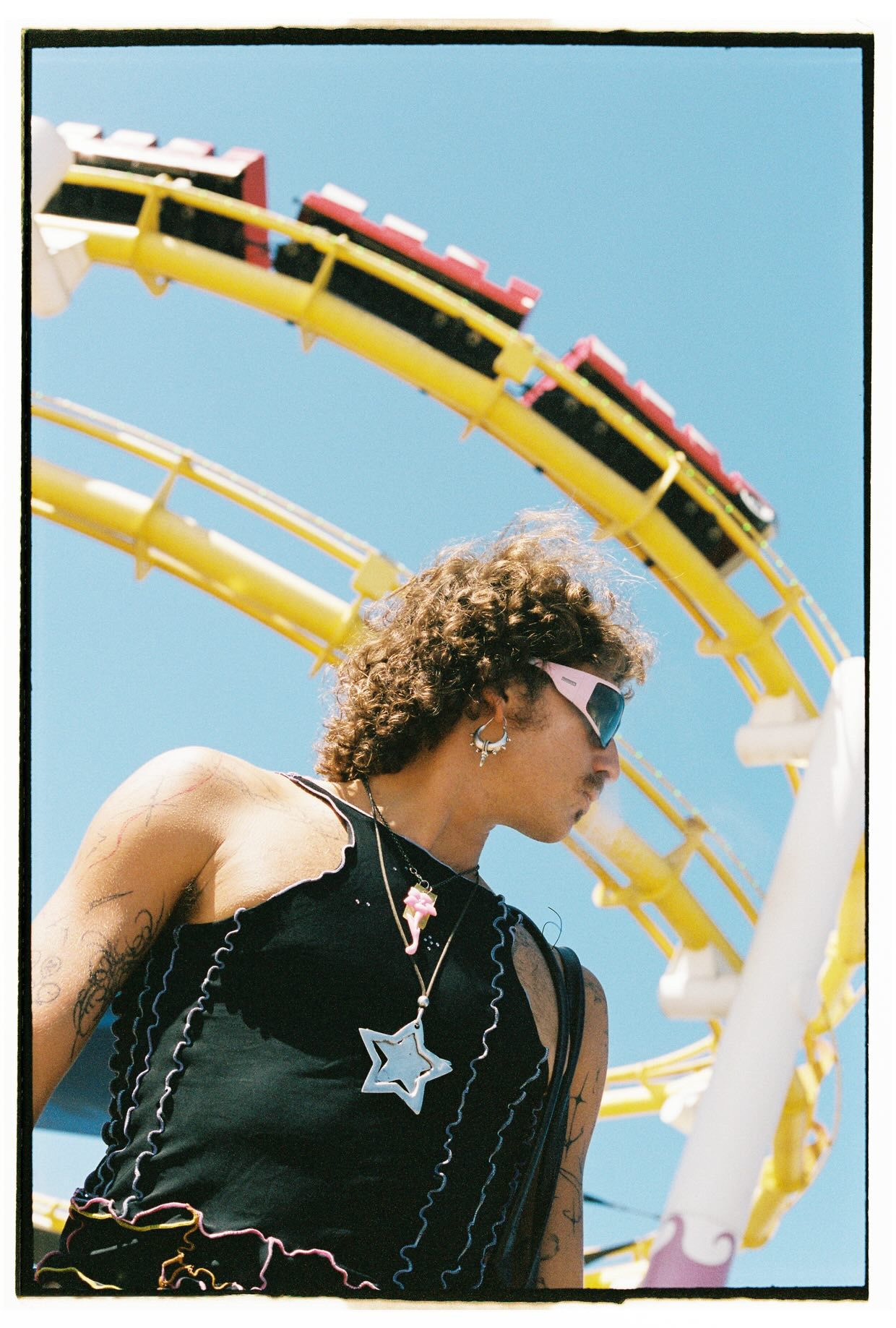 Curly-haired person in black top with star pendant standing before yellow roller coaster track.