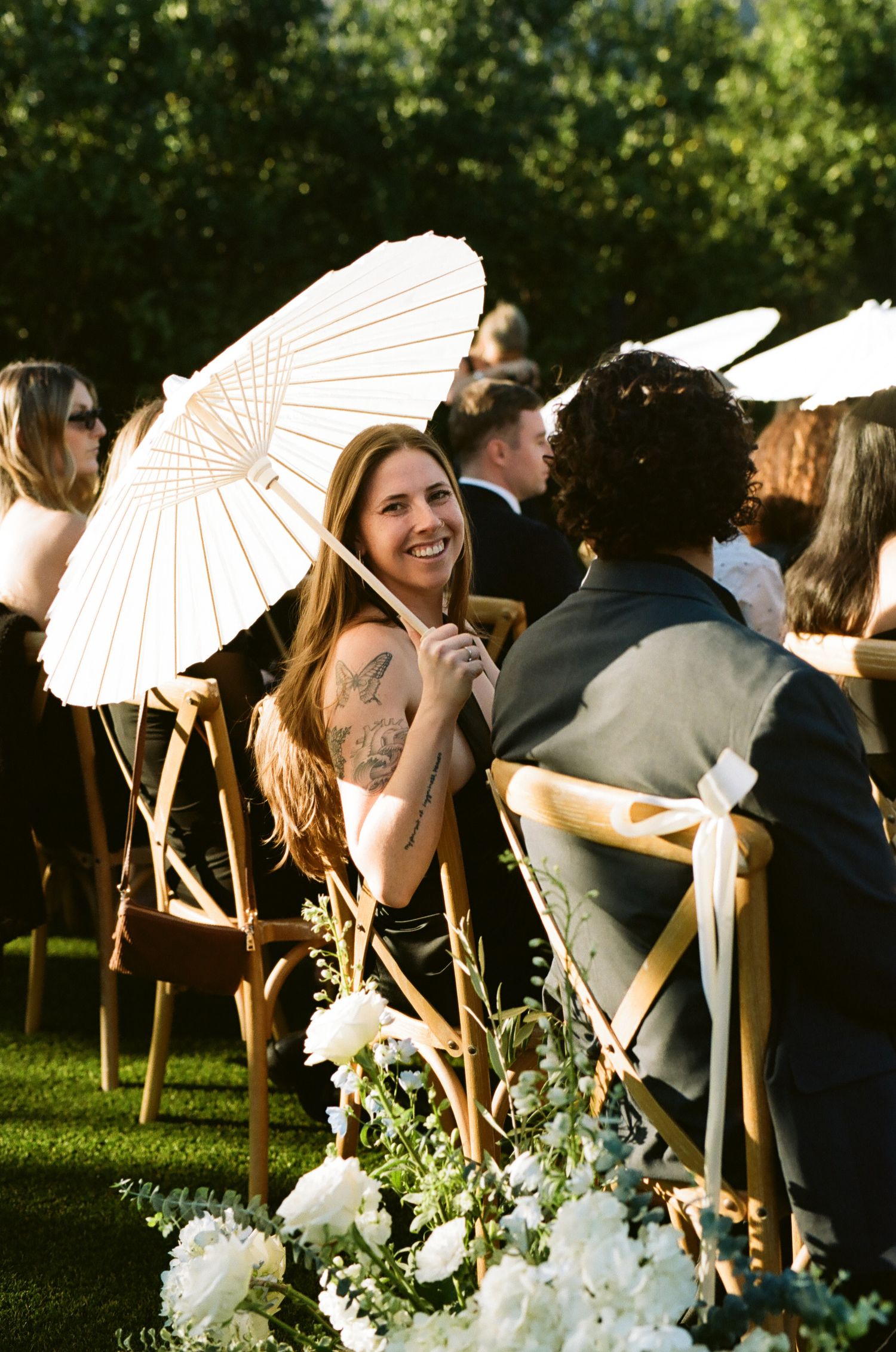 Smiling wedding guest with tattoo holds white parasol at outdoor garden ceremony in wooden chair.