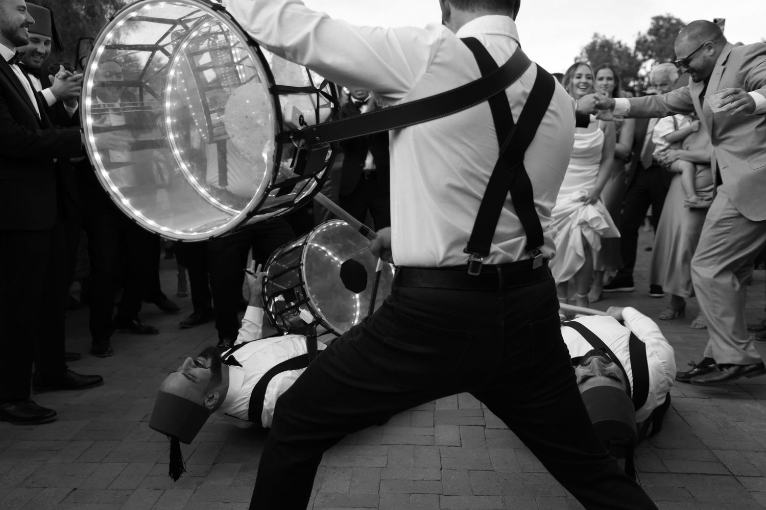 Drummer in suspenders performs energetically with illuminated drum at outdoor wedding celebration.