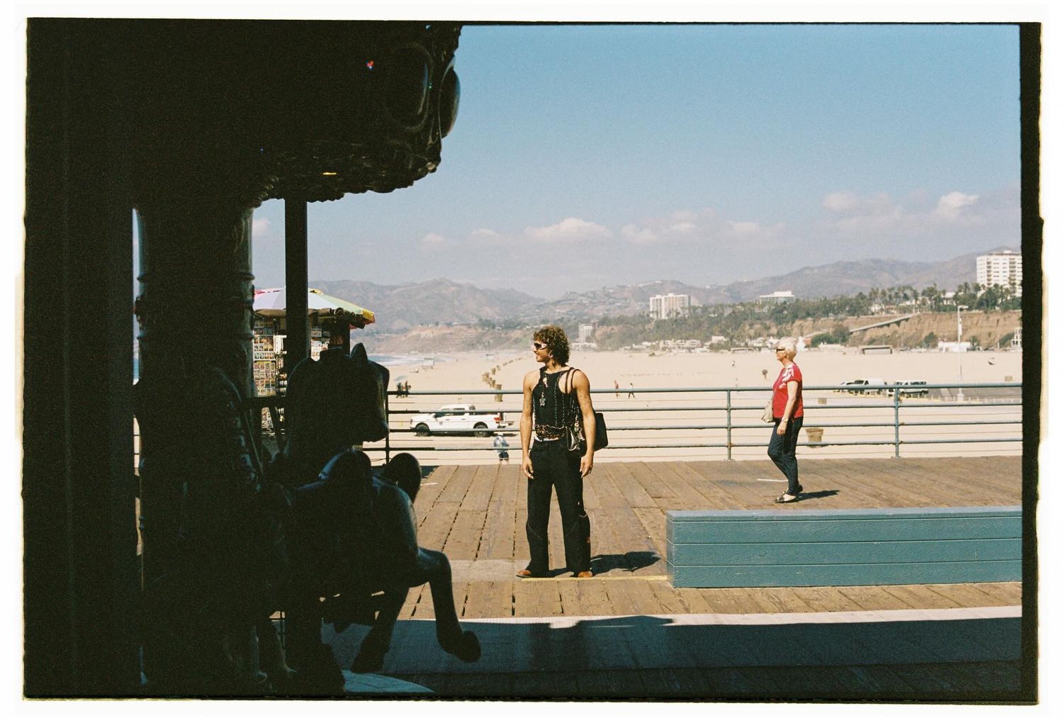 People on sunny boardwalk with mountain and beach views visible in the background.