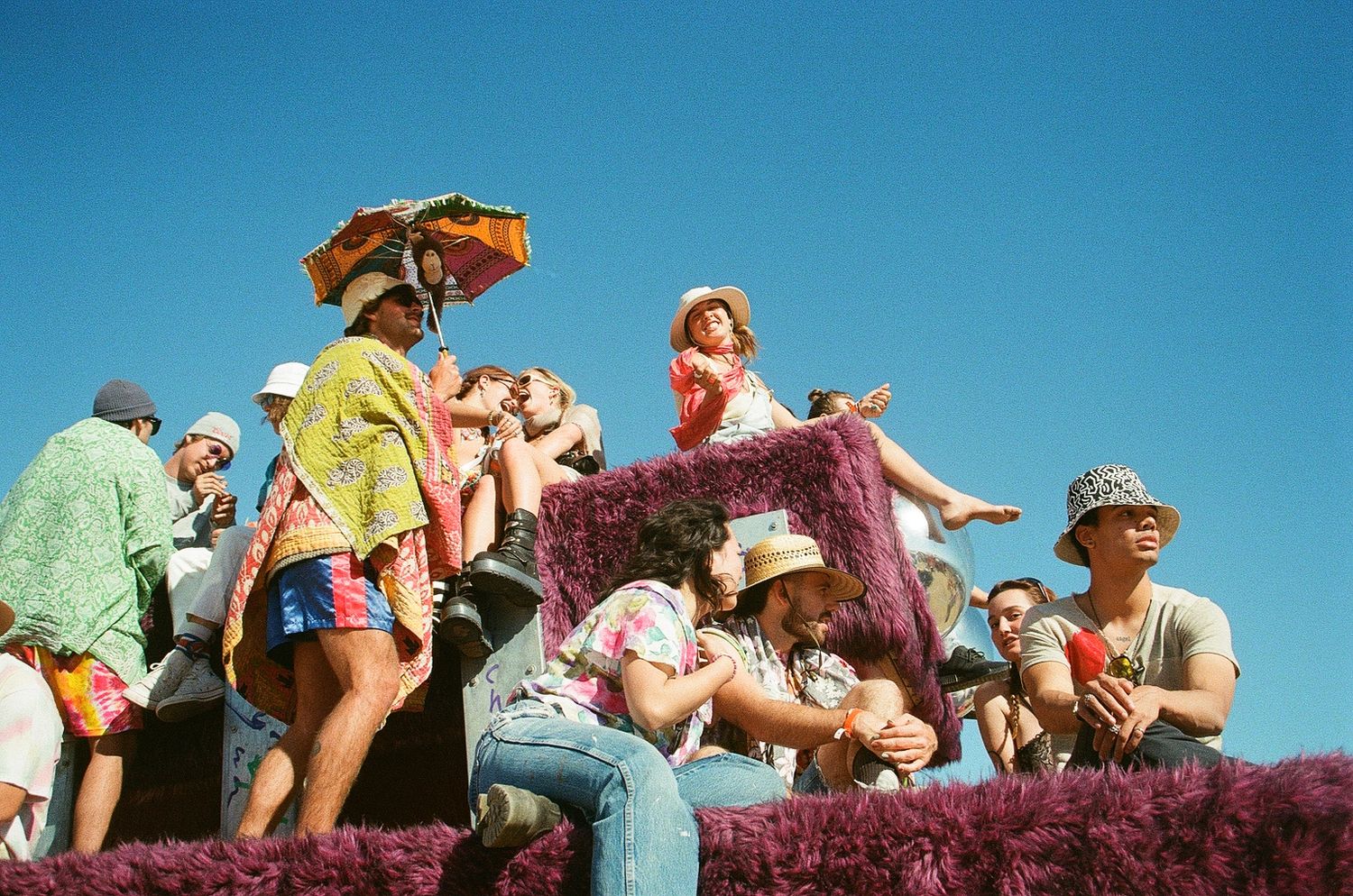 Festival goers in colorful costumes sitting on a float under a clear blue sky.
