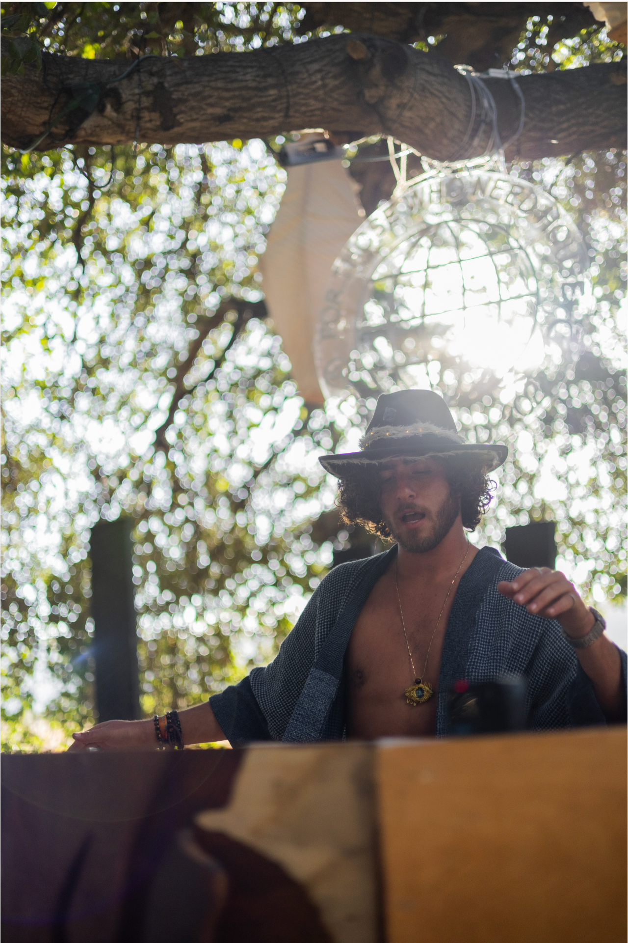 Bohemian man in wide-brim hat and open jacket relaxing under sun-dappled trees at outdoor wedding.