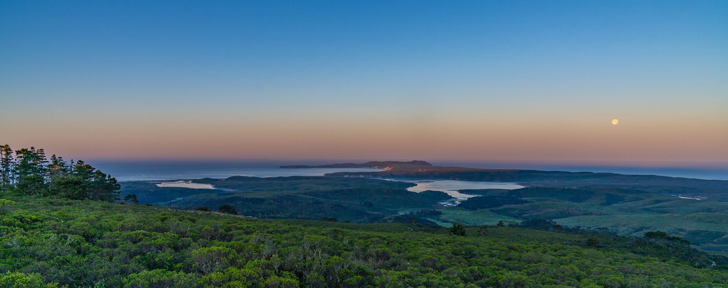 Panoramic twilight view from a hilltop overlooking misty valleys, a lake, and a full moon rising over the horizon.