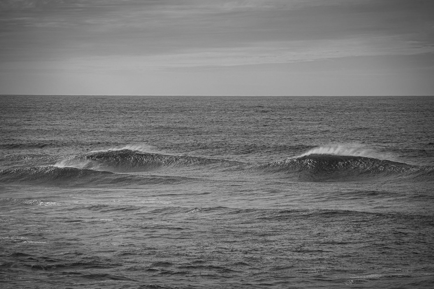 Black and white seascape with gentle ocean waves rolling under a calm overcast sky.
