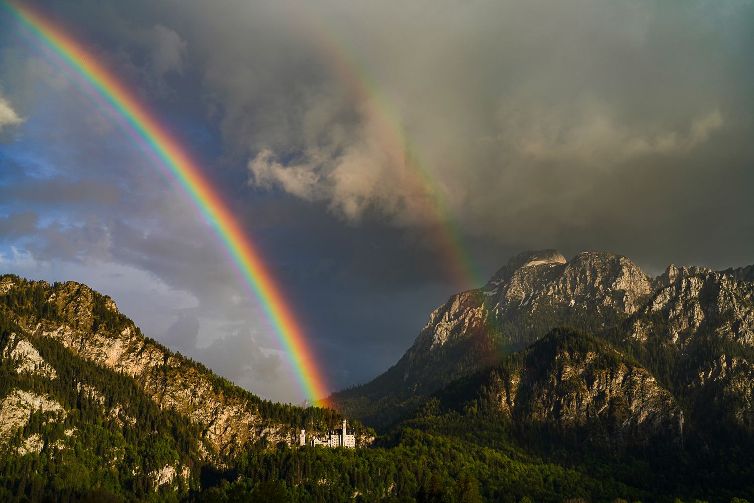 Double rainbow over Neuschwanstein Castle surrounded by dramatic mountain peaks and stormy clouds in Bavaria.