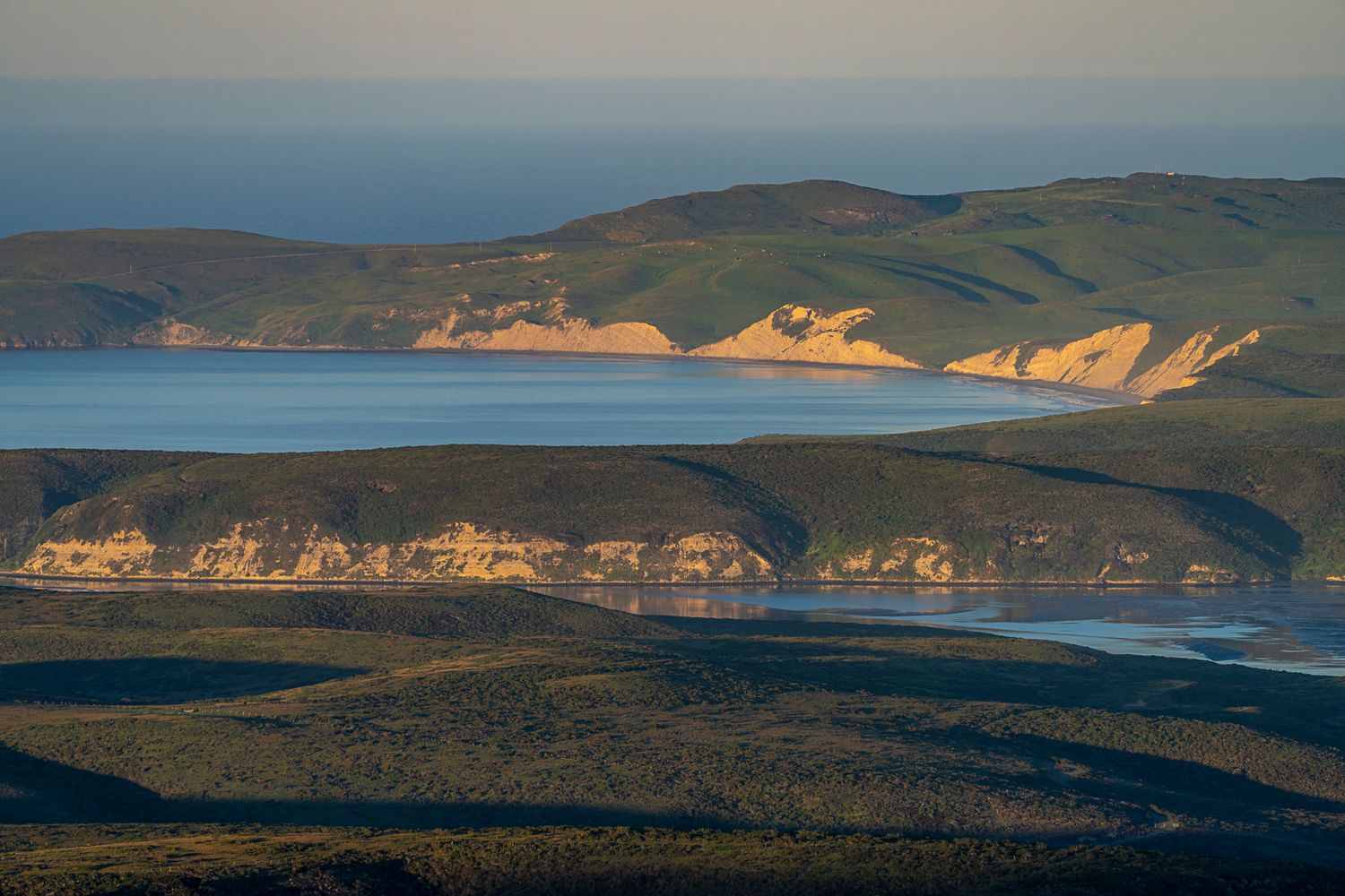 Aerial coastal landscape view of rolling green hills meeting sandy cliffs and blue ocean.
