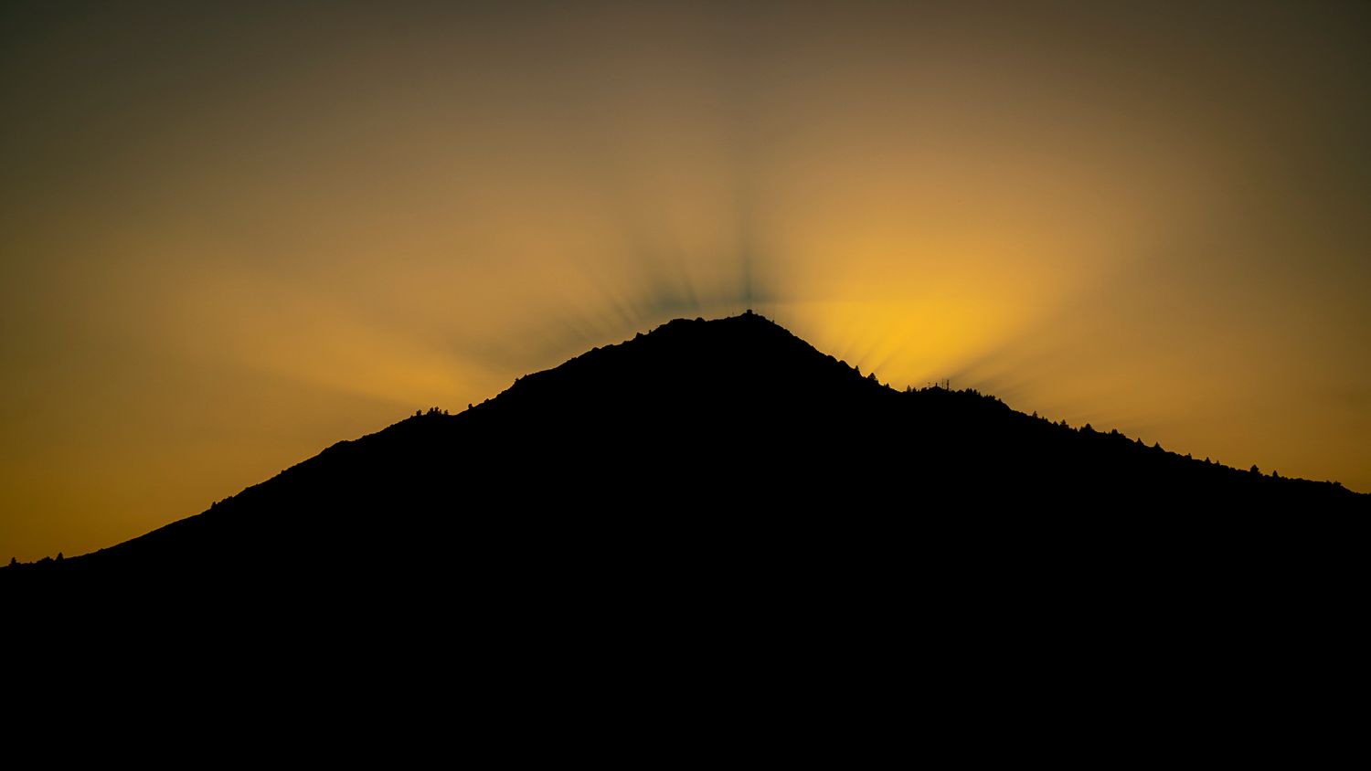 Silhouette of a hill with sun rays bursting from the summit against a golden sky.