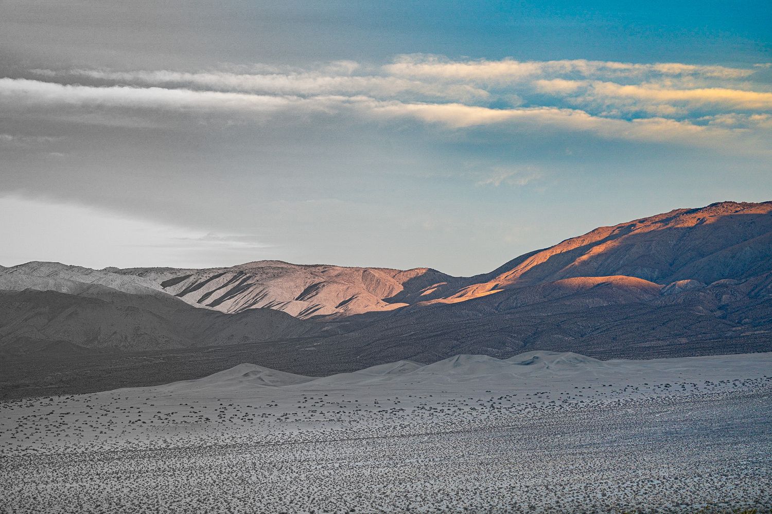 Dramatic desert landscape at sunset with layered mountains and sweeping clouds over a vast arid plain.