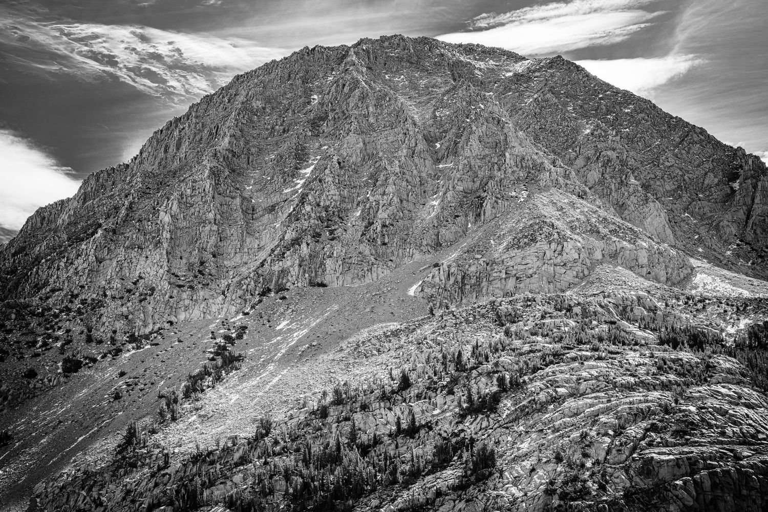 Black and white dramatic rocky mountain peak with sparse trees under a cloudy sky.