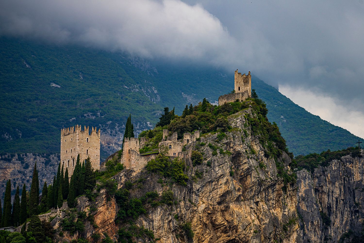 Medieval castle perched dramatically on rocky cliffs with cypress trees and stormy mountain clouds in northern Italy.