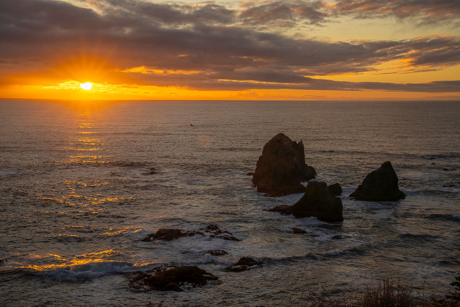 Stunning sunset over rocky Pacific coastline with sea stacks silhouetted against vibrant orange sky.