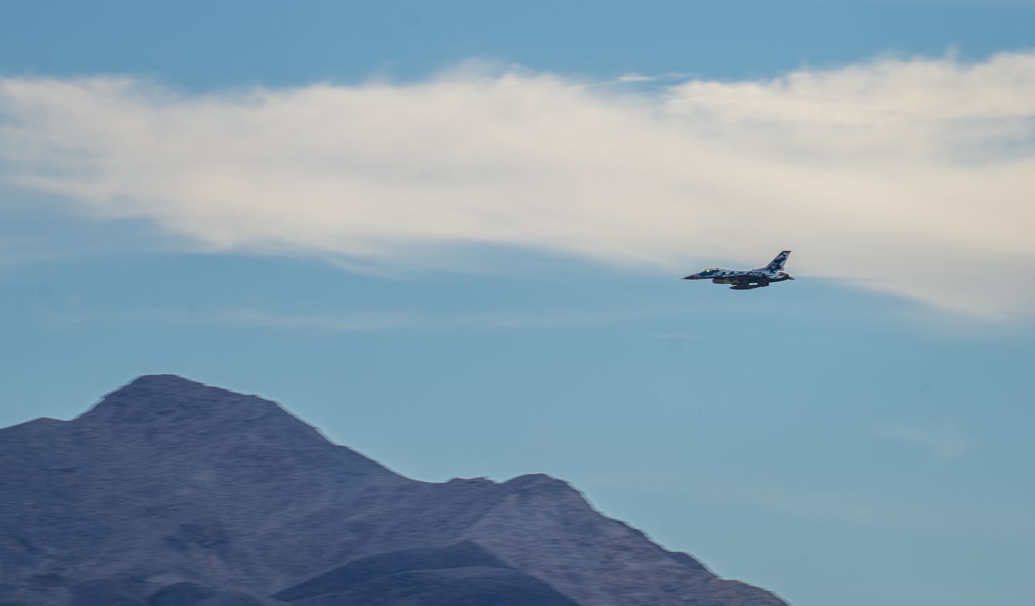 Military jet aircraft flies low over rugged desert mountain peaks against a pale blue sky.