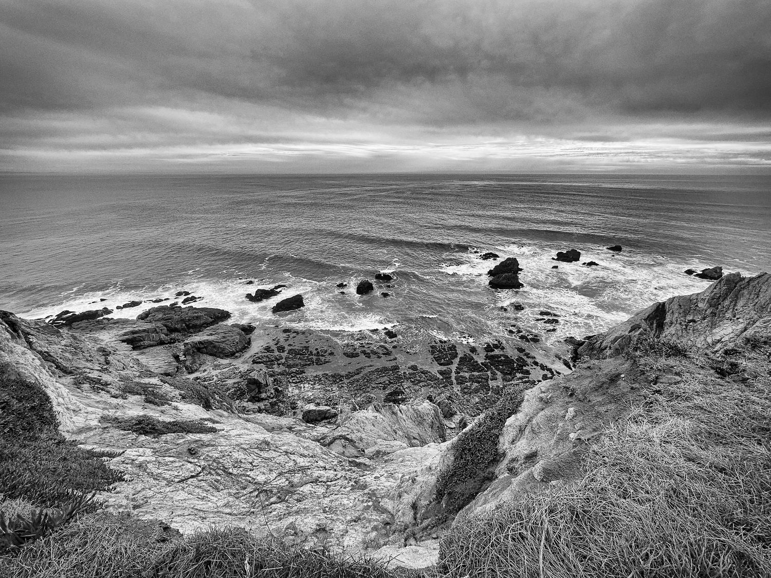 Black and white landscape of eroded coastal cliffs overlooking rocky shoreline with waves under stormy sky.