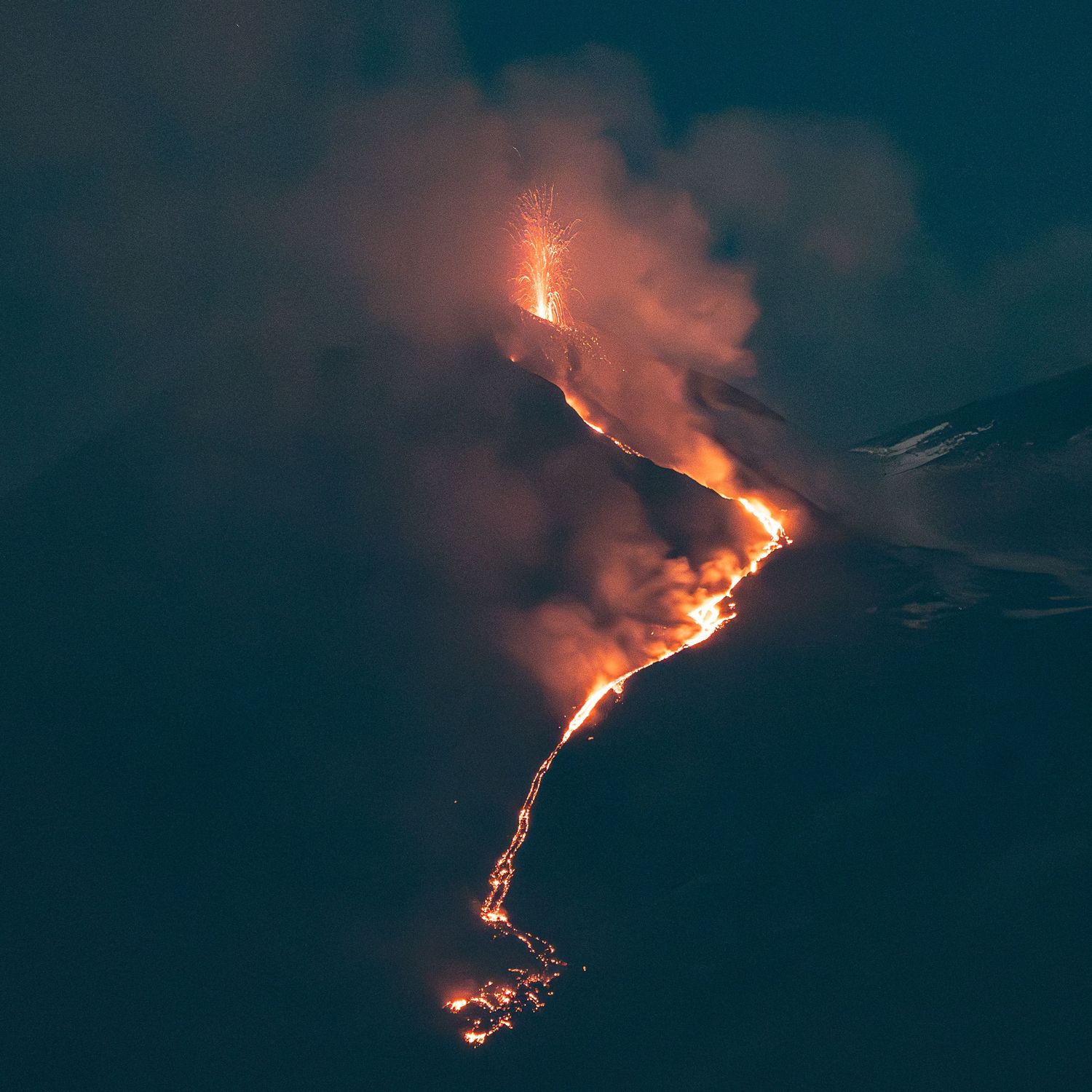 Active volcano erupting at night with glowing lava flows streaming down dark mountainside.