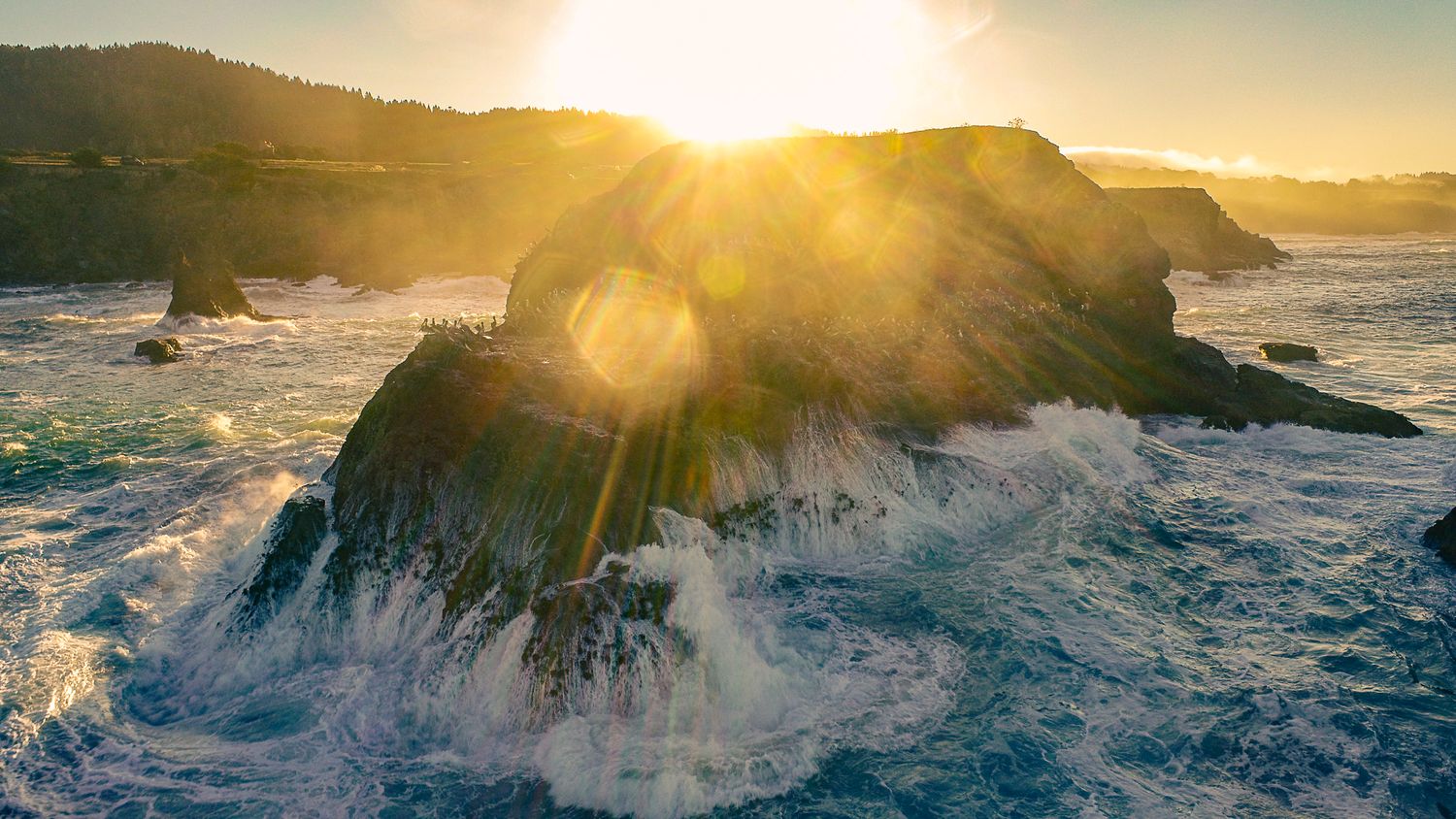 Aerial view of a large ocean rock formation with crashing waves at golden sunset.
