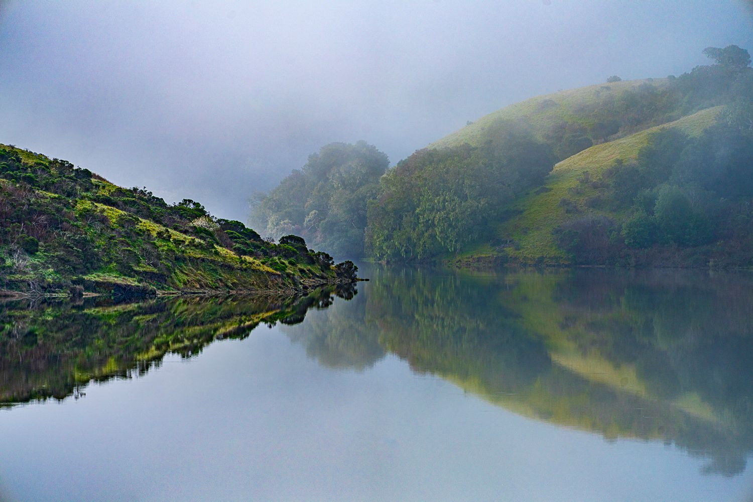 Misty morning fog over a tranquil lake with lush green hills perfectly reflected in water.