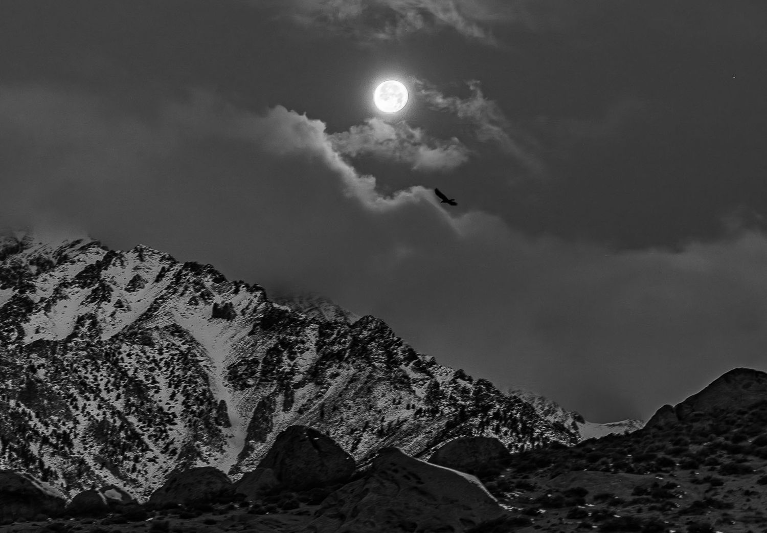Full moon illuminates snow-capped mountain peaks through dramatic storm clouds in monochrome.