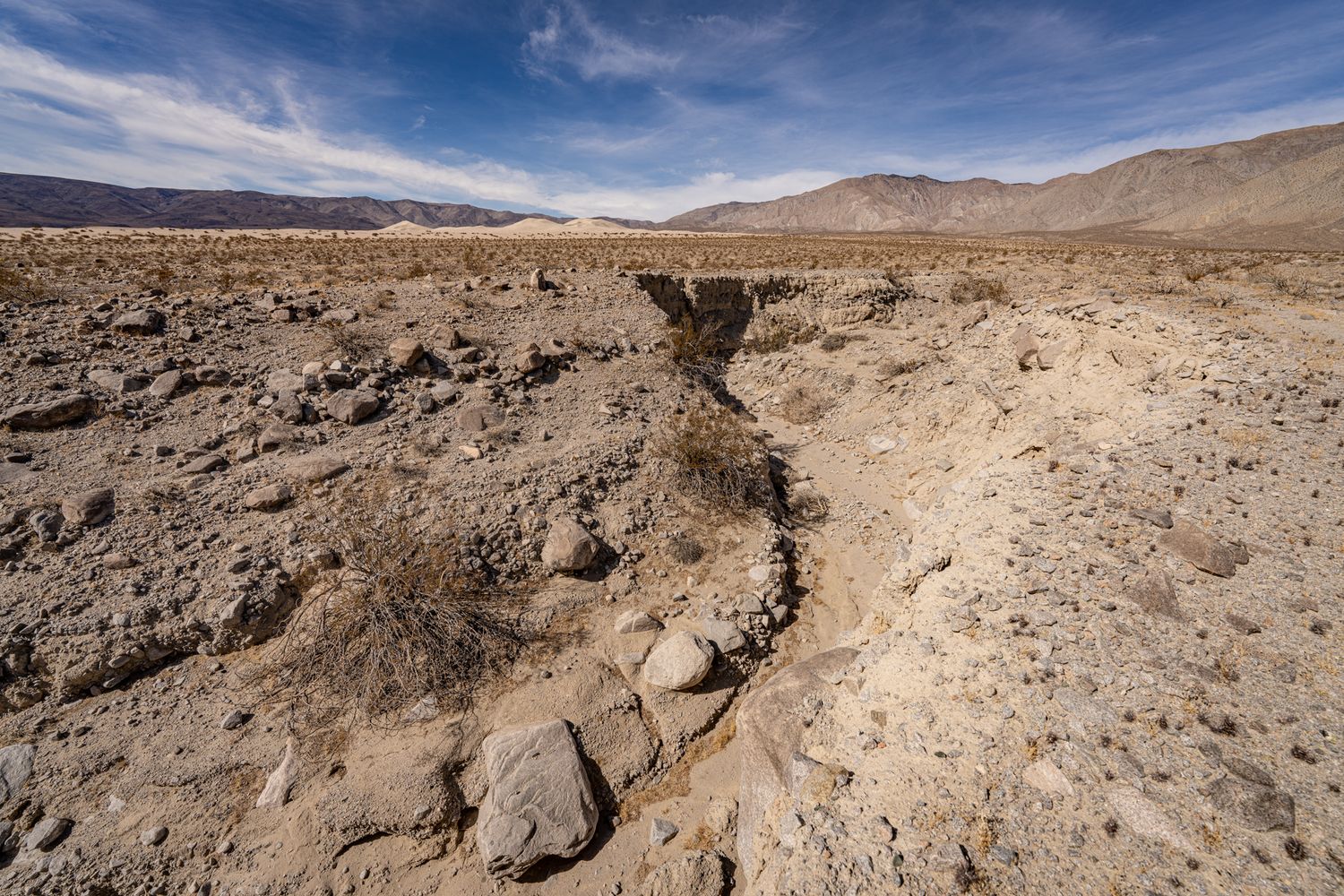 A dry desert wash with eroded rocky terrain and distant mountains under a blue sky.