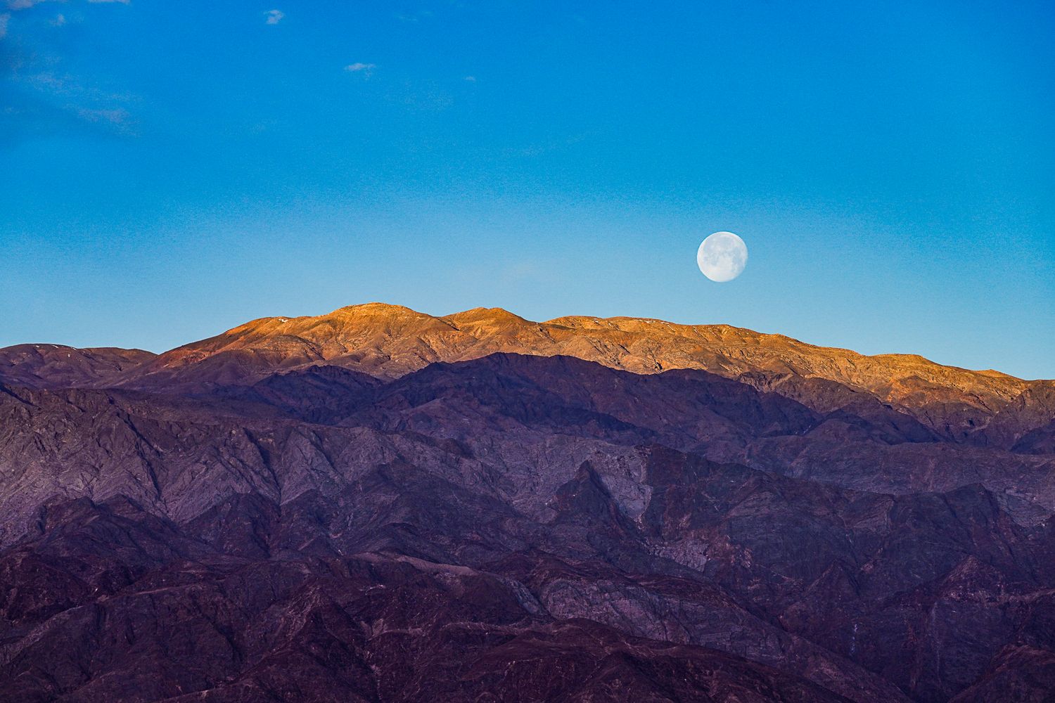 Full moon rises over sunlit desert mountain range against a vivid blue sky.