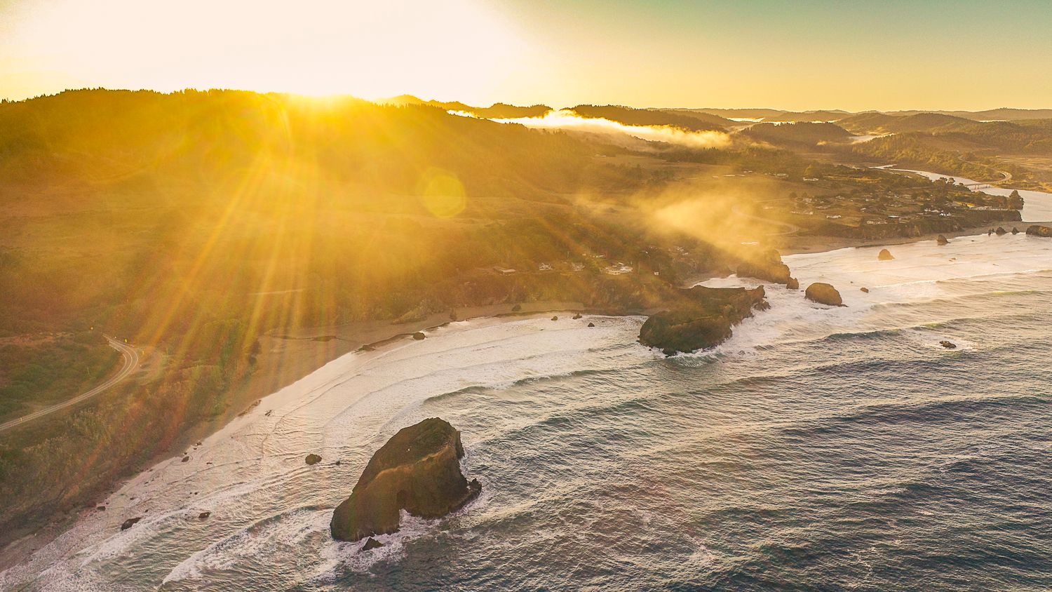 Aerial sunset view of a rugged coastline with golden sunrays illuminating misty ocean waves.