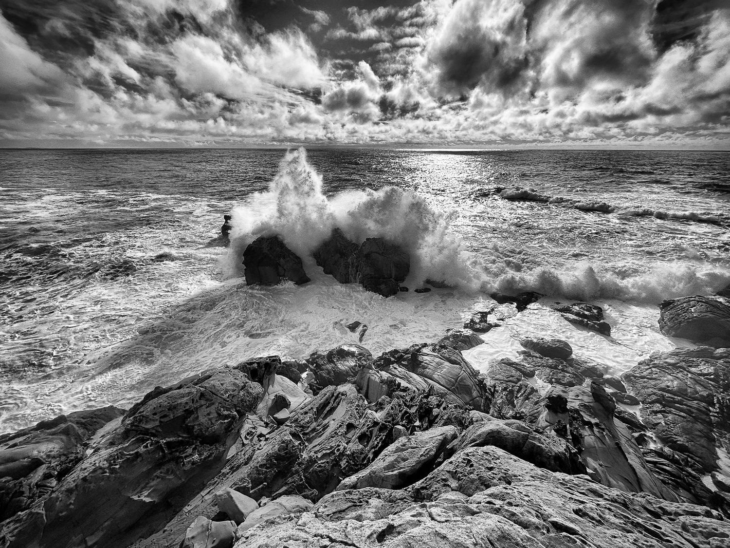 Black and white photo of powerful ocean waves crashing against rocky shore under dramatic cloudy sky.