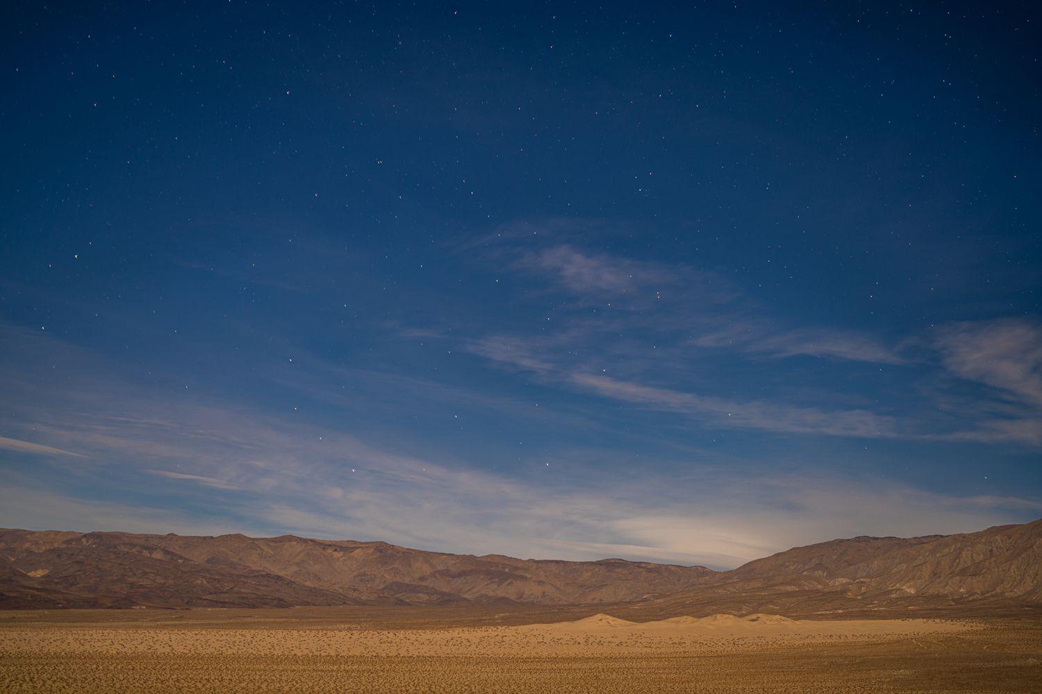 Wide desert landscape at night with wispy clouds, starry blue sky, and mountain range on the horizon.