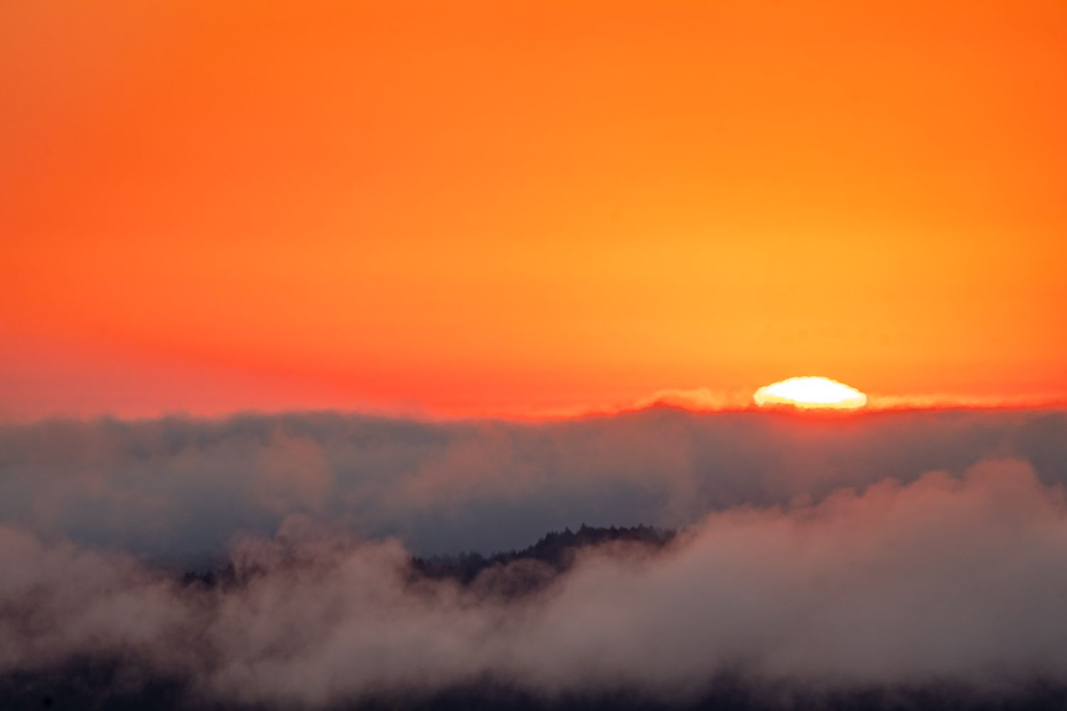 Vibrant orange sunset with the sun setting behind a layer of clouds over misty mountain peaks.