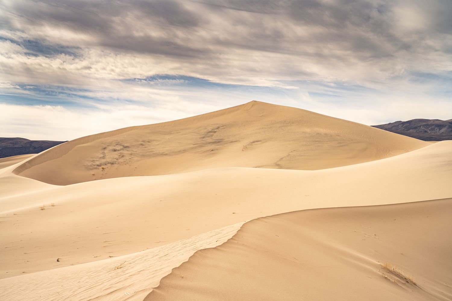 Large smooth sand dune rises against a cloudy sky with distant mountains in a vast desert landscape.