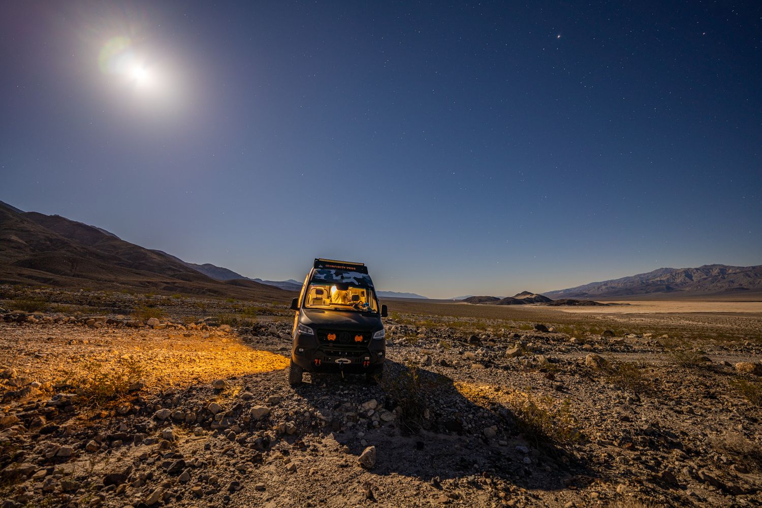 Adventure van parked in moonlit desert landscape at night with mountains visible in the background.