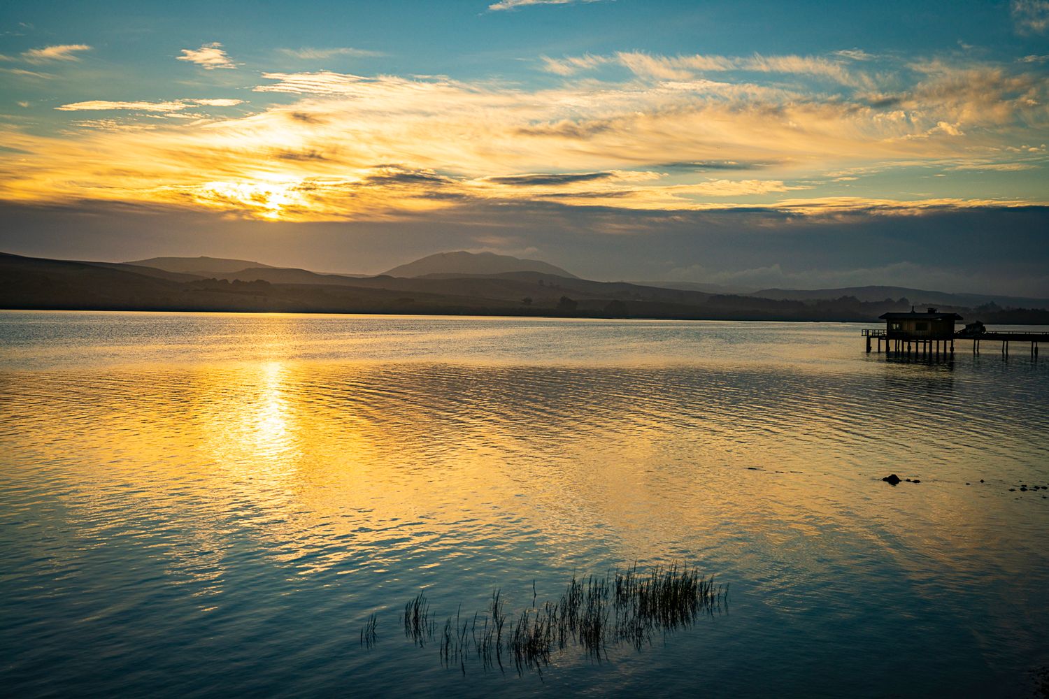Golden sunset reflecting on a calm lake with mountain silhouettes and a distant pier.