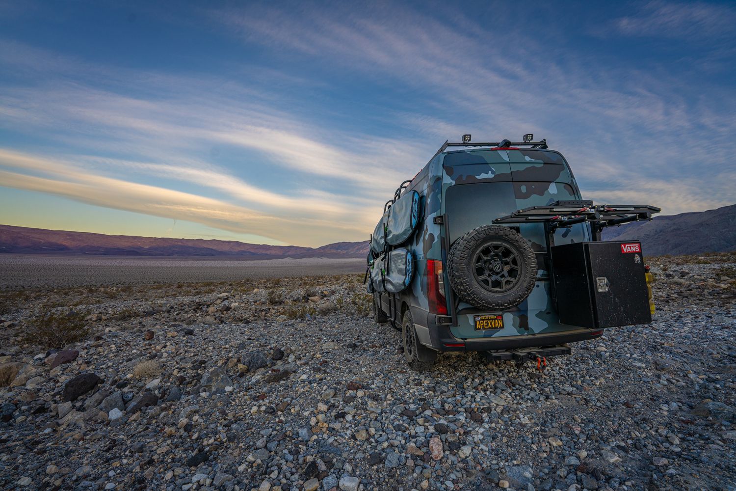 A rugged off-road van parked on rocky desert terrain at sunset with mountains in the background.