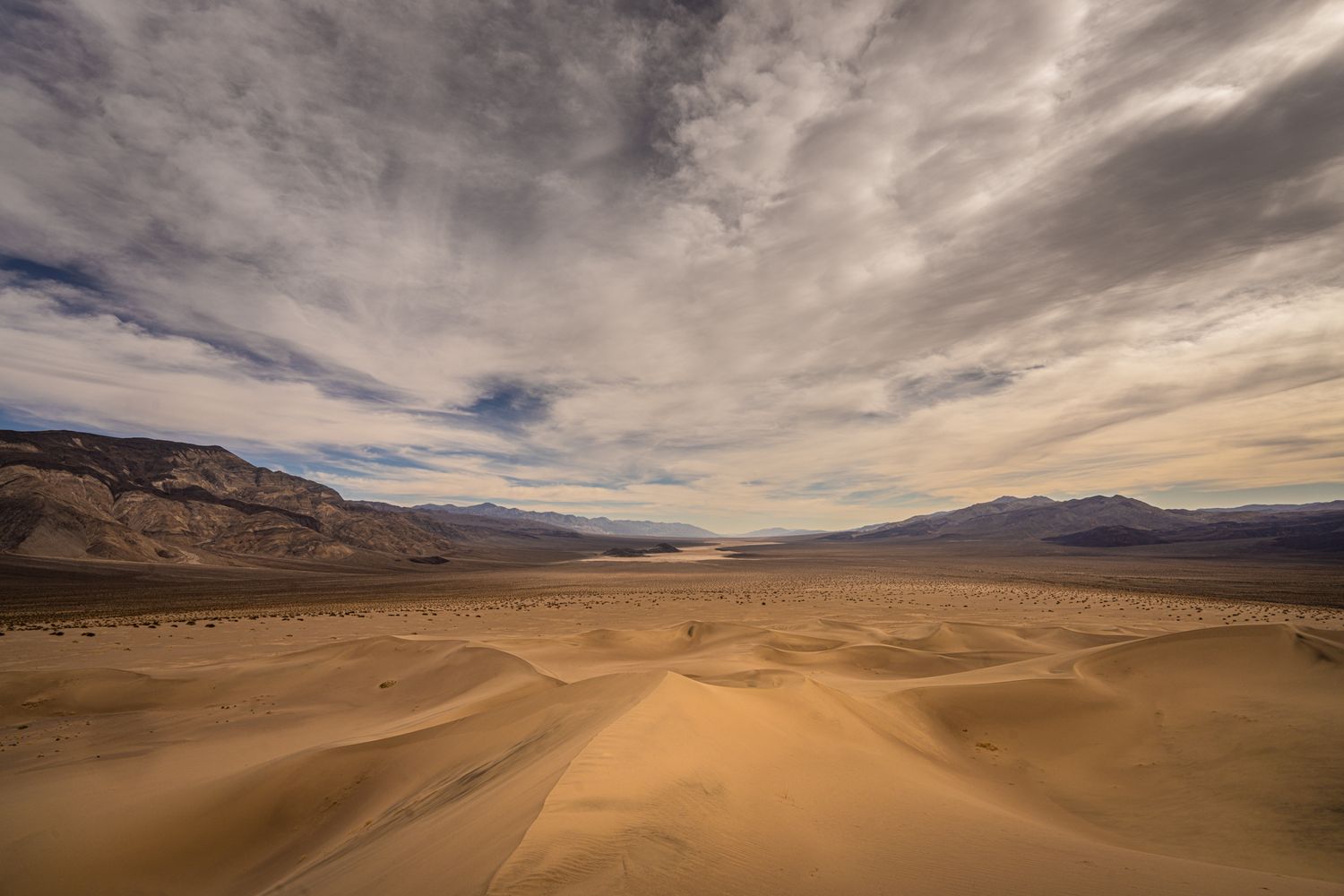 Vast golden sand dunes stretch toward distant mountains under dramatic cloudy skies in a desert landscape.