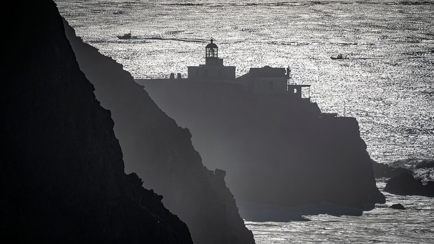 Black and white photo of a lighthouse perched on dramatic coastal cliffs above crashing ocean waves.