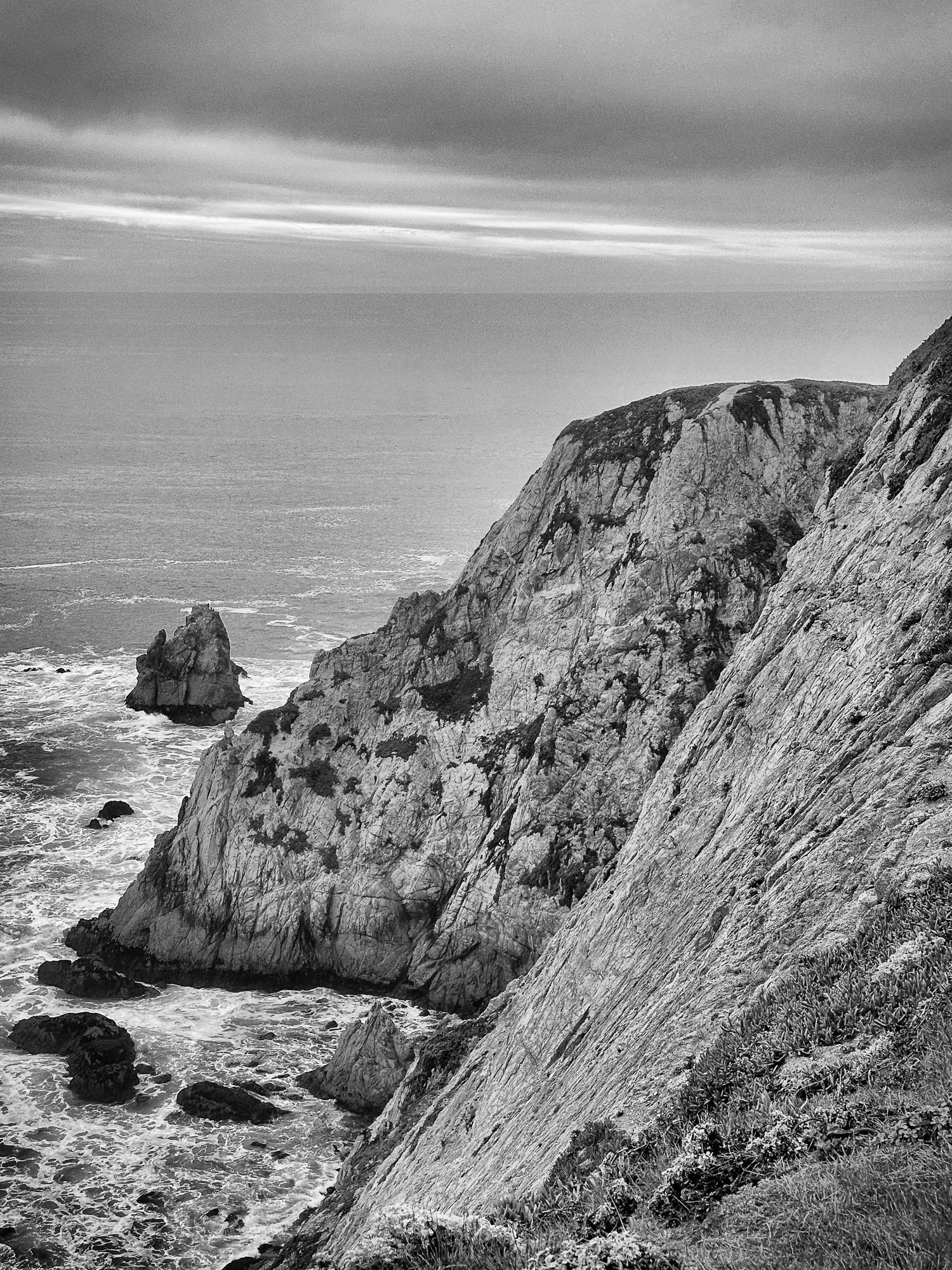 Dramatic black and white photo of rugged sea cliffs with rock formations rising from churning ocean waters.