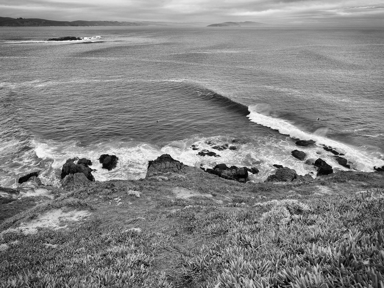 Black and white aerial view of rocky Pacific coastline with crashing waves and vast ocean horizon.