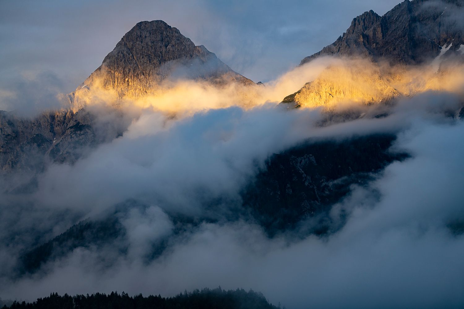 Golden sunrise light illuminating rugged mountain peaks dramatically emerging through thick clouds.