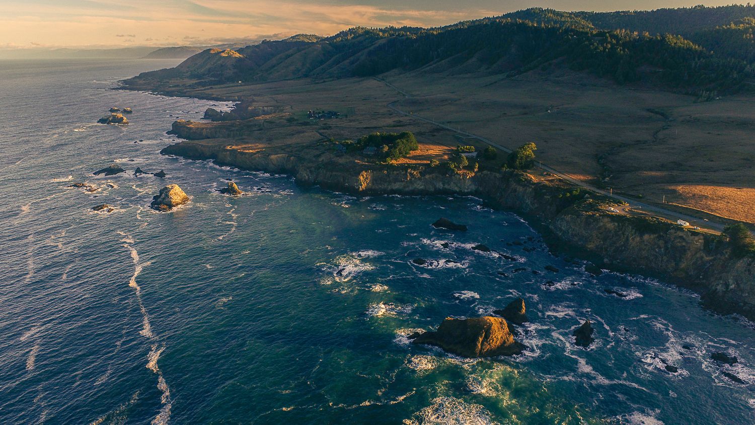 Aerial sunset view of a dramatic rocky coastline with ocean waves and rolling green hills.