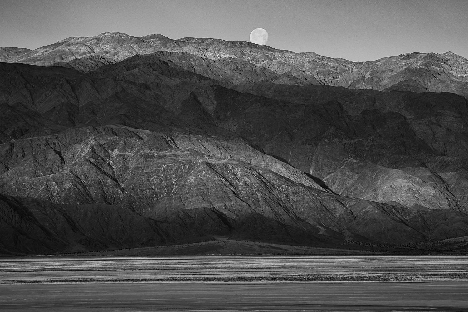 Black and white photograph of a full moon rising over rugged mountain ridgeline above a desert basin.