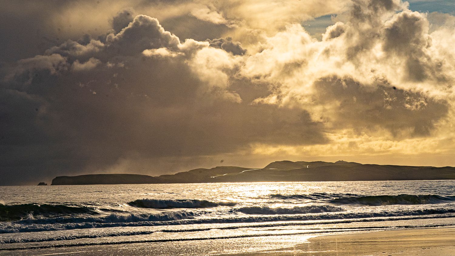 Golden sunset light reflects on ocean waves at a sandy beach with stormy clouds.