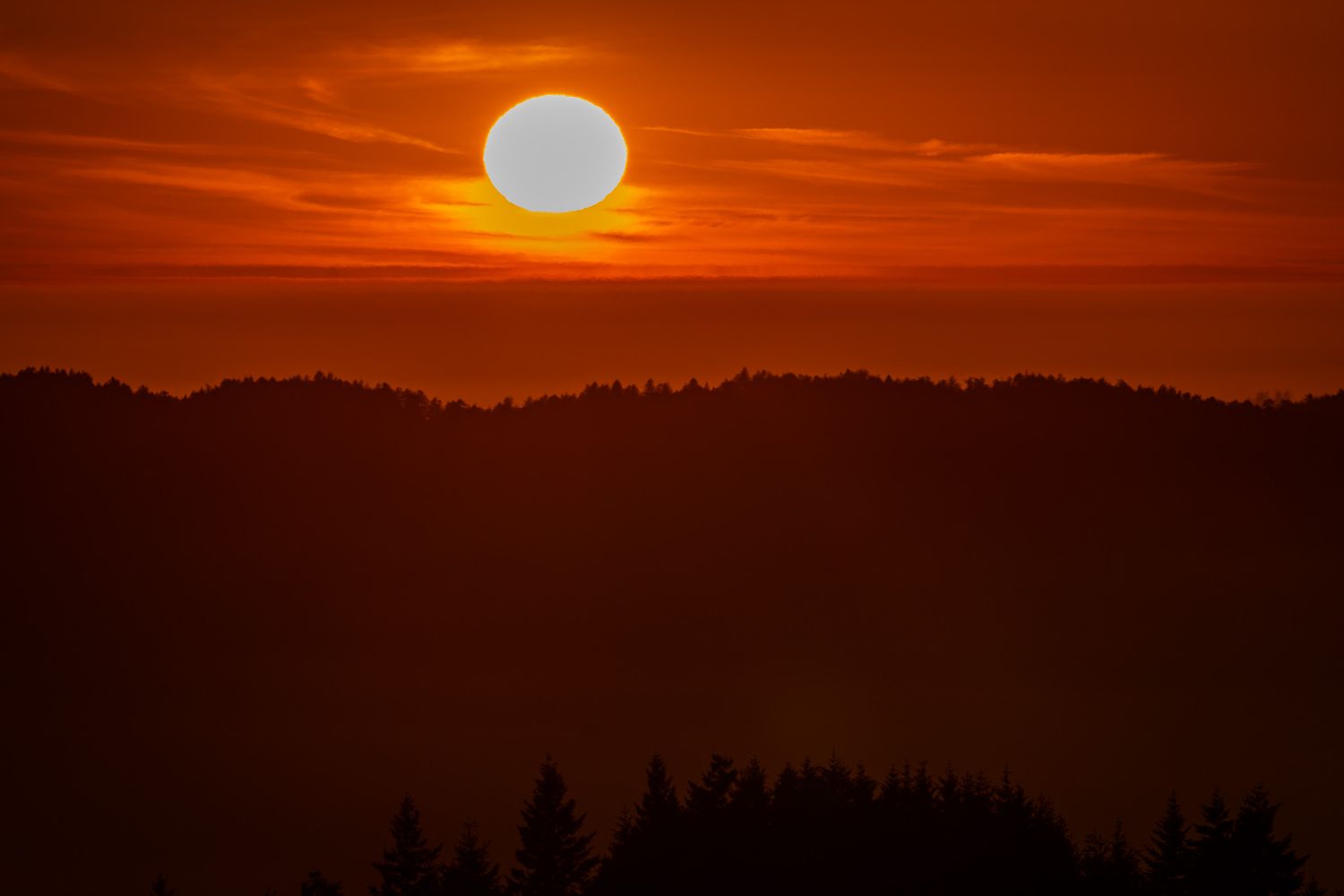 Dramatic red and orange sunset over silhouetted forest hills with glowing white sun against vibrant sky.