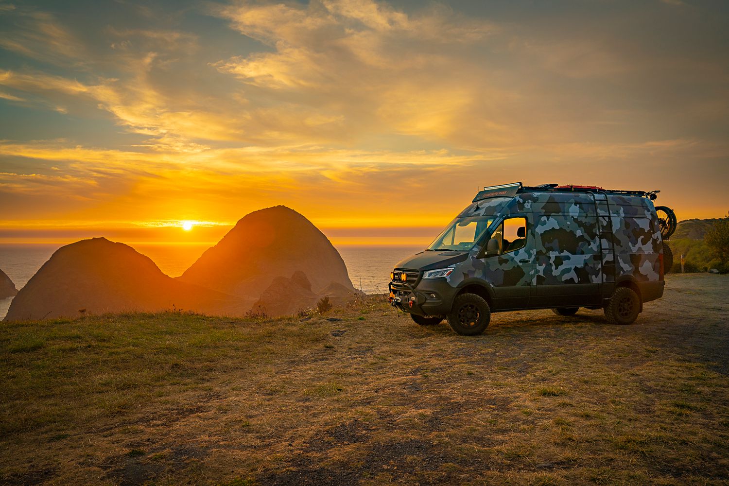 A camouflage-painted adventure van parked on a coastal bluff during a vibrant golden Oregon sunset.