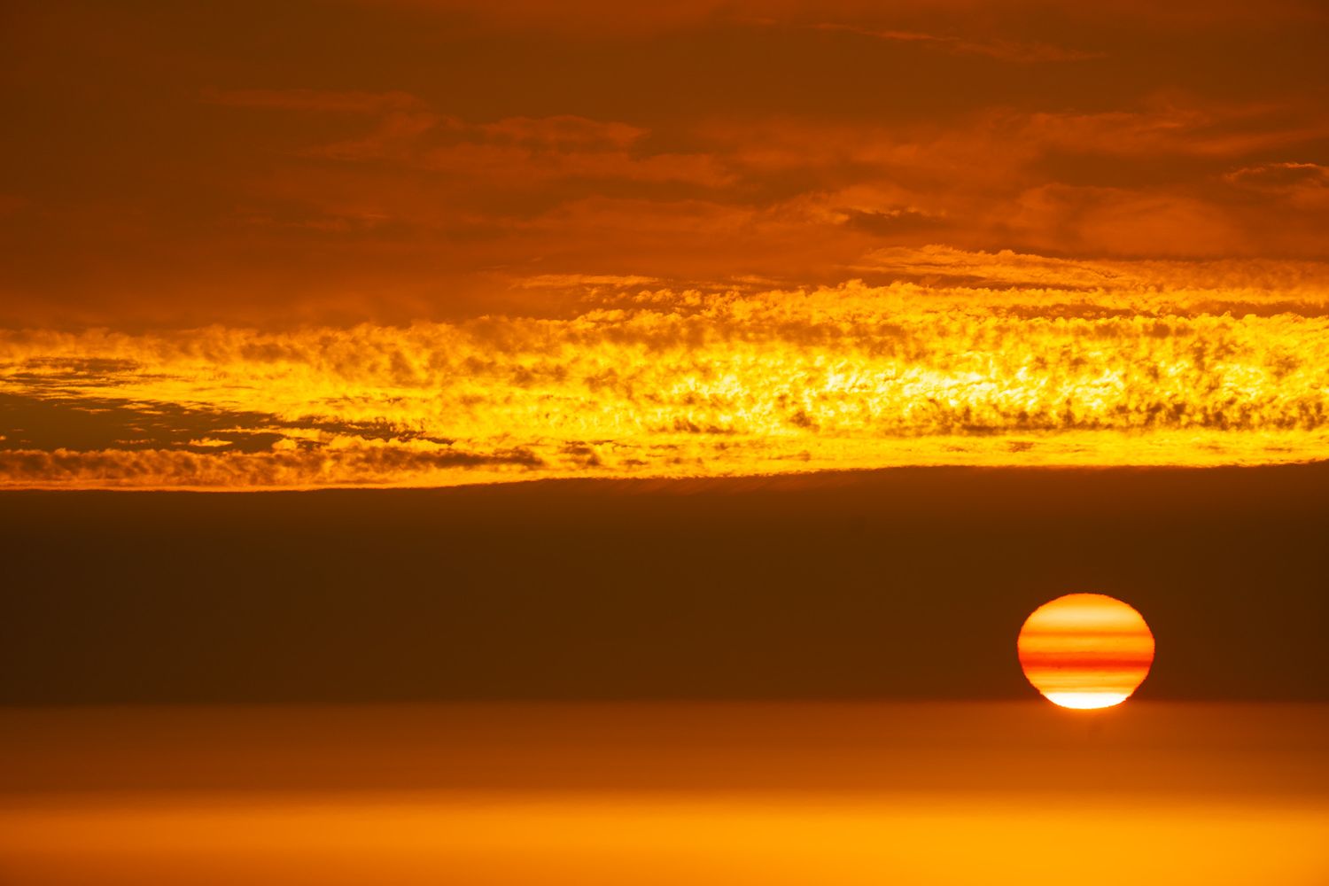 Golden sunset over the ocean with vibrant orange sky, dramatic clouds, and a striped sun on the horizon.