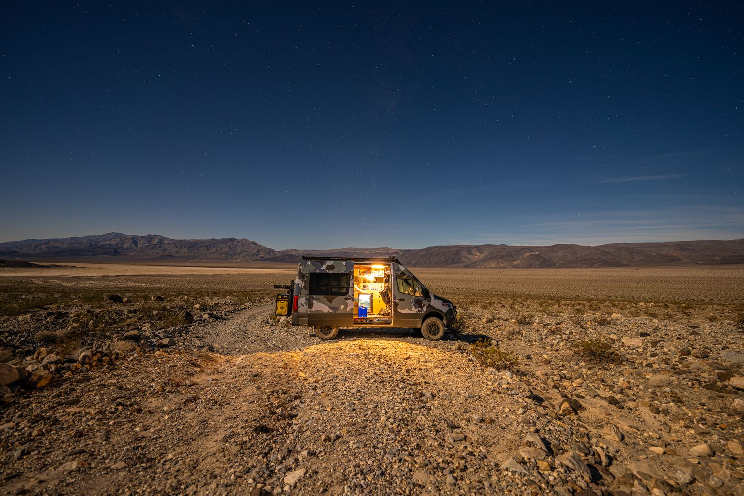 Camper van illuminated at night in vast desert landscape under starry blue sky with distant mountains.