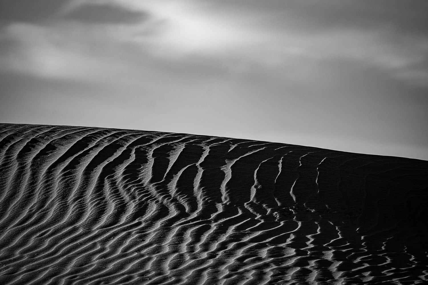 Minimalist black and white photography of rippling sand dune patterns under a cloudy sky.