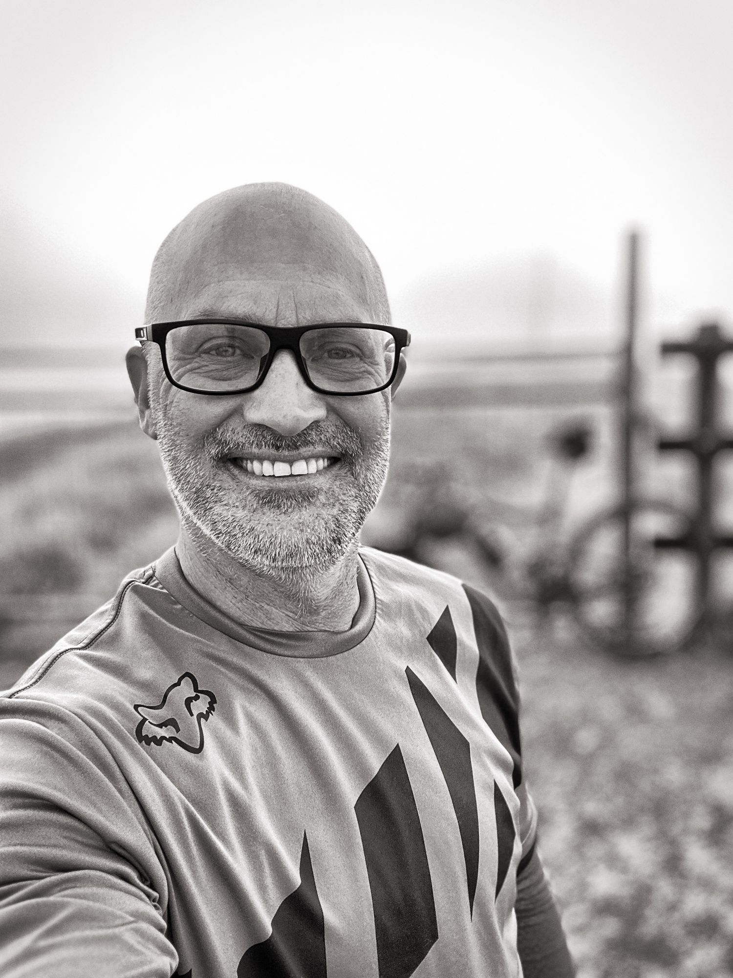 Bald man with glasses smiling in a black and white selfie outdoors with a bicycle visible in the foggy background.
