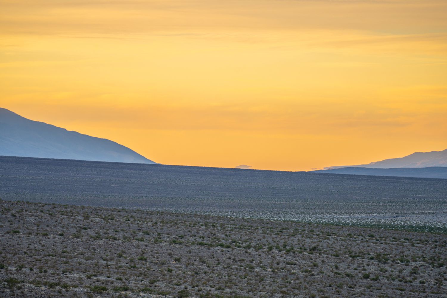 Golden orange sunset sky over flat desert plains with layered mountain silhouettes in the distance.